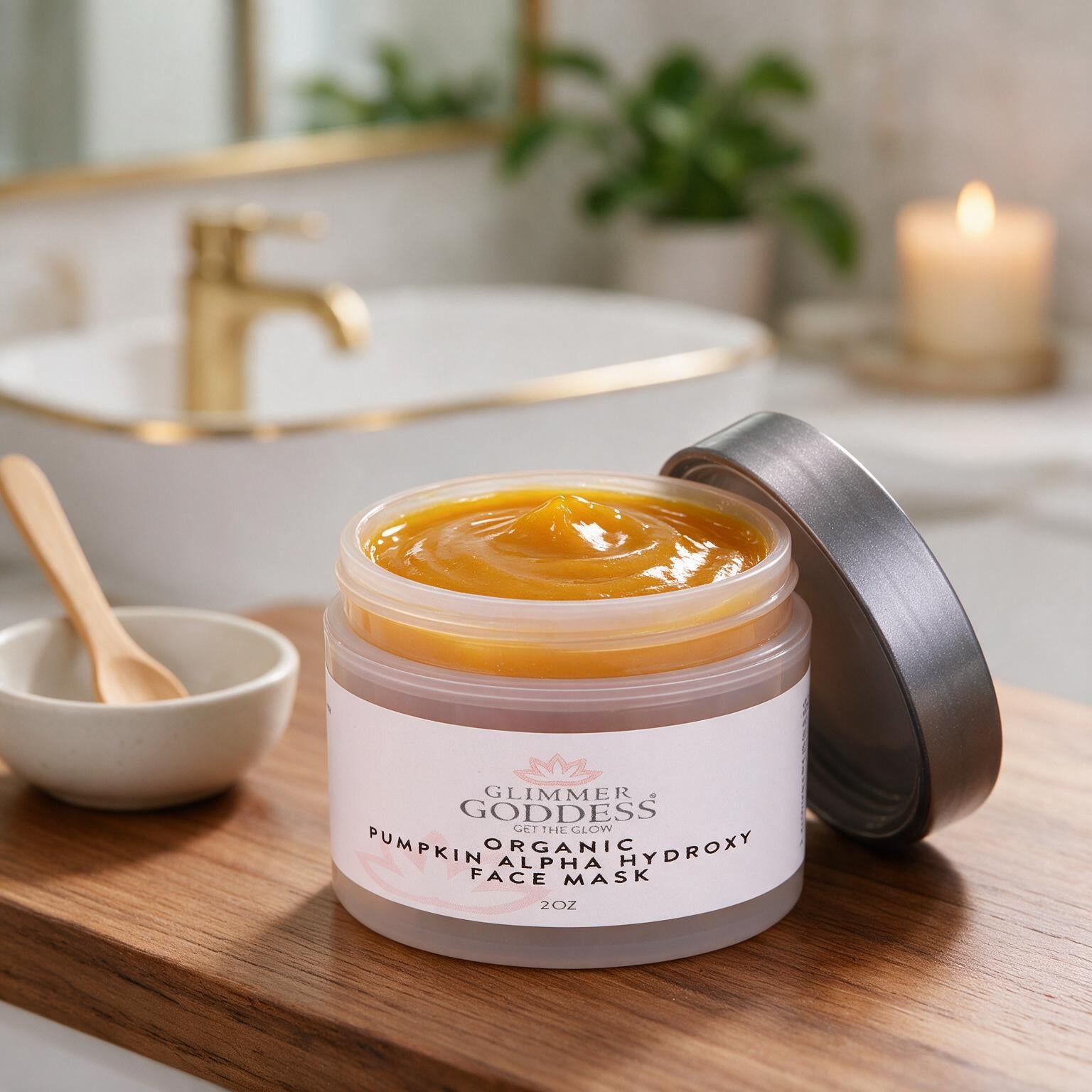 An open jar of Glimmer Goddess Organic Pumpkin Alpha Hydroxy Face Mask sits on a wooden tray in a softly blurred bathroom setting. The mask is a rich golden orange, with its silver lid leaning beside it.