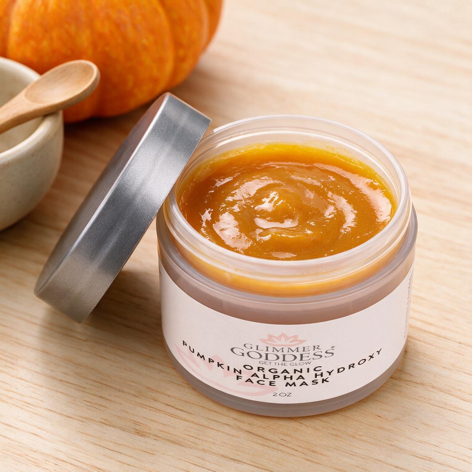 An open jar of Summer of Goddesses Organic Alpha Hydroxy Face Mask with pumpkin scent, showing the orange-yellow mask inside, with its silver lid resting beside it on a light wood surface.