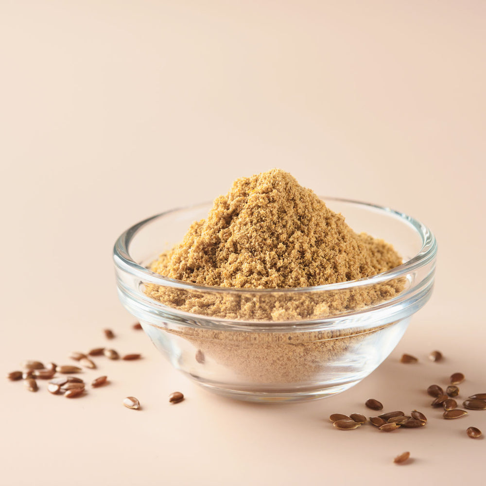 Glass bowl of ground flaxseed with whole flax seeds on a beige background